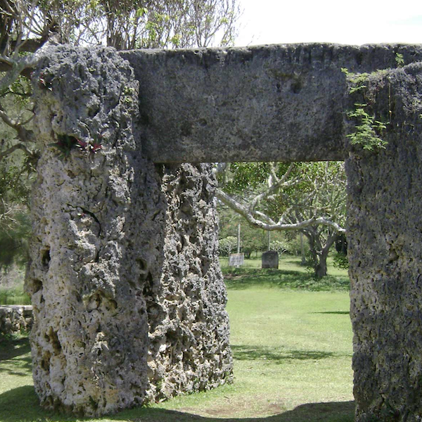 A stone monument with two stones acting as posts and a third stone sitting vertically atop the others. The monument sits on grass and there are trees seen in the background. 