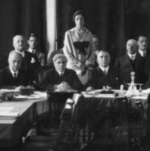 Thumbnail image of a meeting of the League of Nations. It shows three men sitting at the head of the table with other four men and a woman standing behind them.